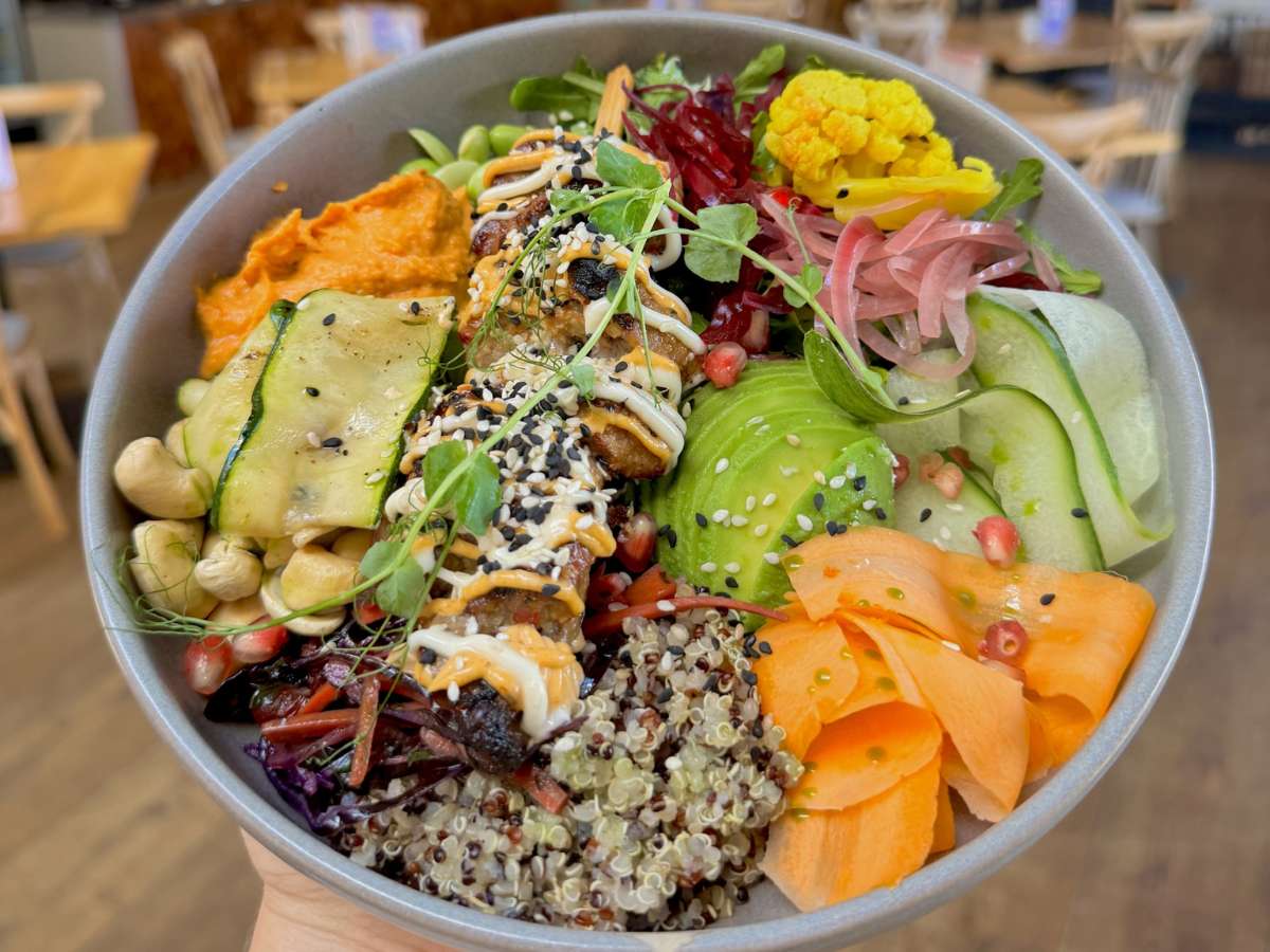 A colourful nourish bowl filled with quinoa, avocado, roasted vegetables, hummus, edamame beans, pickled onions, beetroot, carrot ribbons, cucumber, cashew nuts and sesame-coated plant protein, finished with a creamy drizzle and microgreens. The bowl is being held inside Forage Farm Shop Café with tables softly blurred in the background.