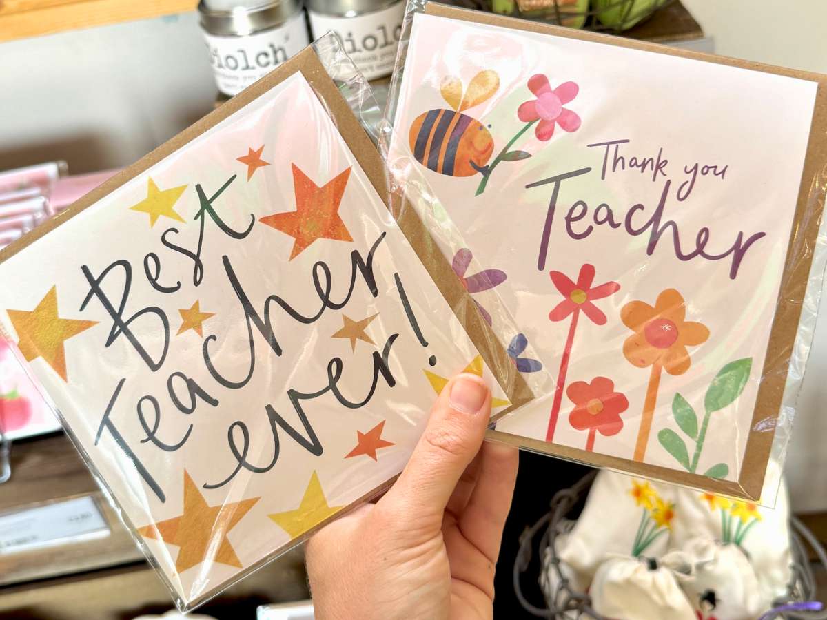 A hand holding two colourful greeting cards from the Forage gift range. One reads "Best Teacher Ever!" with gold stars, while the other says "Thank you Teacher" surrounded by bright flowers and a bee. The cards are displayed inside the farm shop with gifts and homeware softly visible in the background.